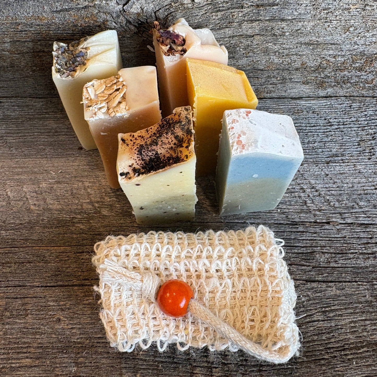 A group of handmade artisan soaps in various colors and textures, arranged on a rustic wooden surface alongside a small cream-colored woven pouch with a red-orange bead closure.