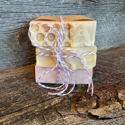 Three stacked bars of handmade soap tied with pink and white twine, set against a rustic wooden background.