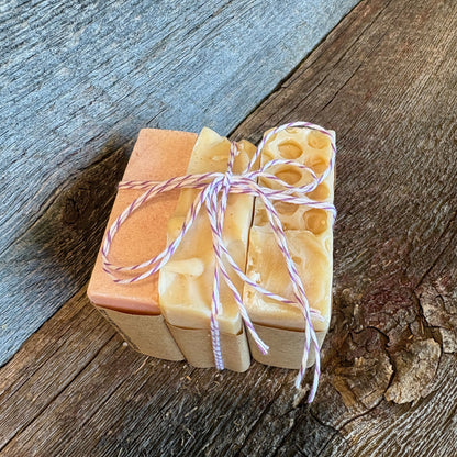 Three stacked bars of handmade soap in shades of cream and light tan, tied with red and white bakers twine, set on a rustic wooden surface.