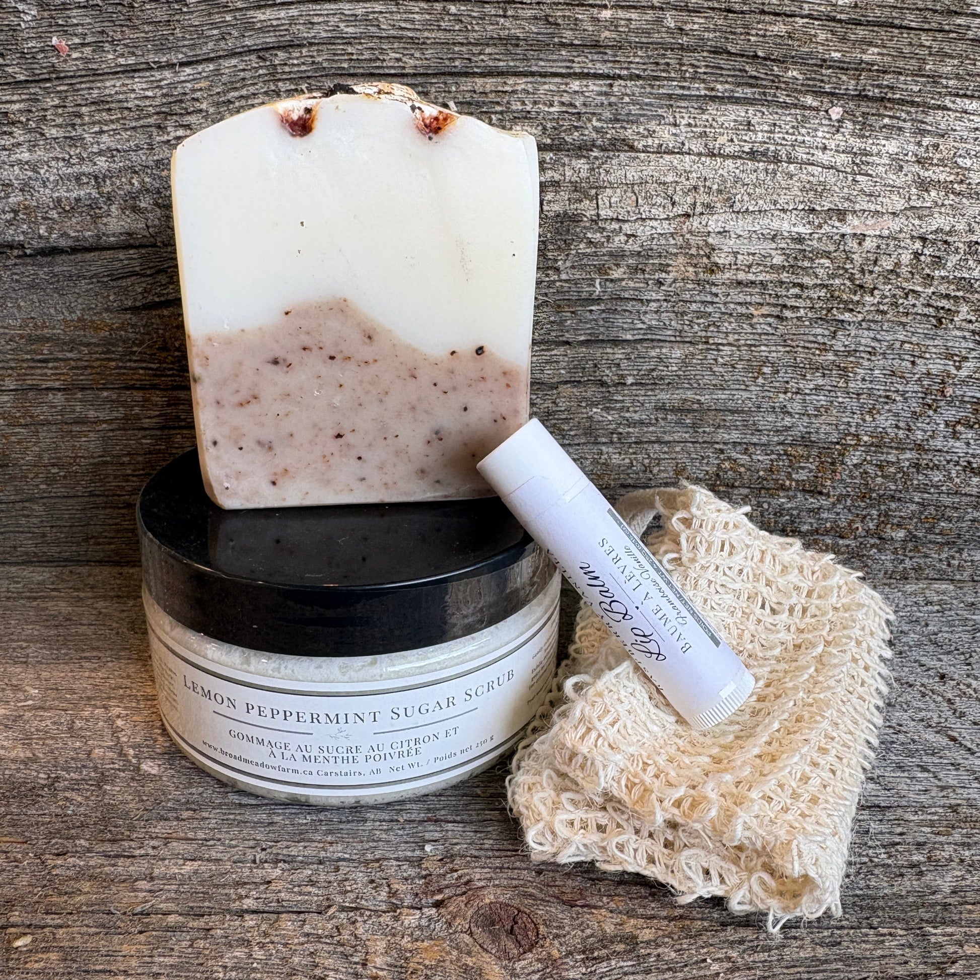 A bar of handmade soap with a white and speckled beige lower section stands on top of a black-lidded jar labeled 'Lemon Peppermint Sugar Scrub.' A white lip balm tube and a beige knitted cleansing cloth rest beside them on a rustic wooden surface.