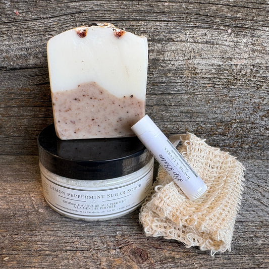 A bar of handmade soap with a white and speckled beige lower section stands on top of a black-lidded jar labeled 'Lemon Peppermint Sugar Scrub.' A white lip balm tube and a beige knitted cleansing cloth rest beside them on a rustic wooden surface.