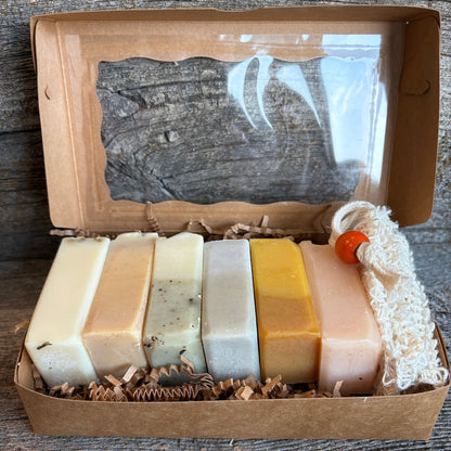 A set of six artisanal handmade soaps in various earthy colors, presented in a brown kraft gift box with a clear window and shredded paper filler, set against a rustic wooden background.