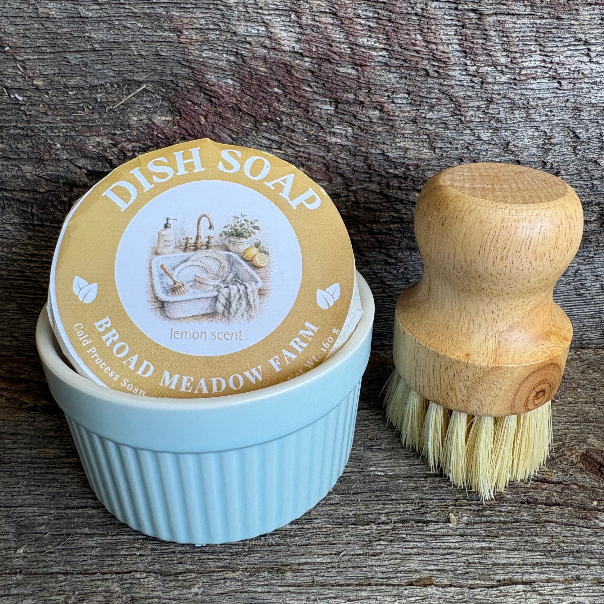 A light blue ribbed dish soap container with a round lid label reading 'DISH SOAP BROAD MEADOW FARM' and 'lemon scent,' shown beside a wooden-handled dish brush on a rustic weathered wood surface.