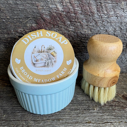 A light blue ribbed dish soap container with a round lid label reading 'DISH SOAP BROAD MEADOW FARM' and 'lemon scent,' shown beside a wooden-handled dish brush on a rustic weathered wood surface.