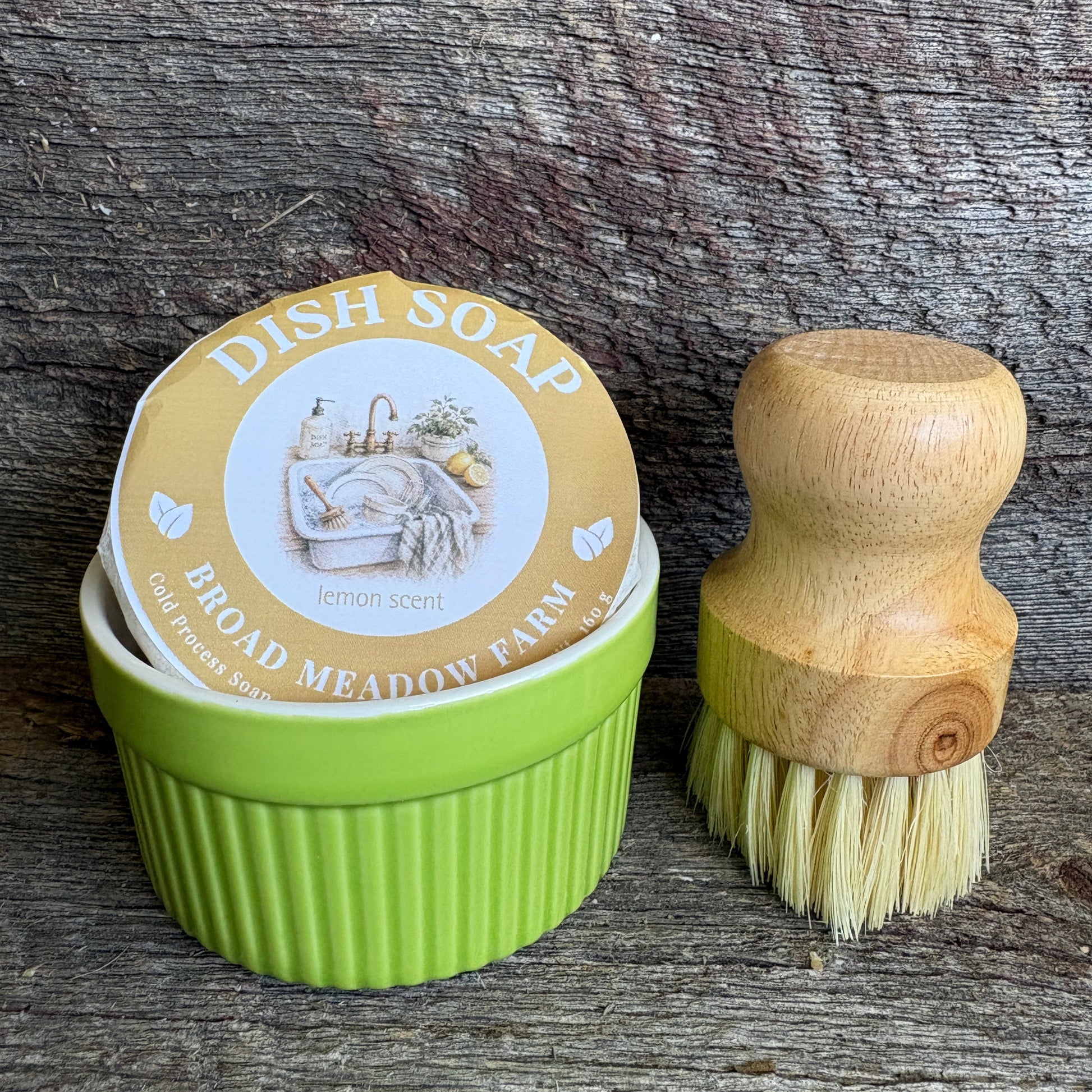 A green ribbed dish soap container with a round lid label reading 'DISH SOAP BROAD MEADOW FARM Lemon Scent,' shown next to a wooden-handled brush with cream bristles on a rustic weathered wood surface.