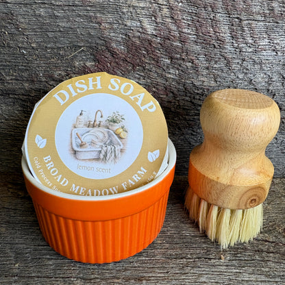 A small orange tub of Broad Meadow Farm lemon-scent dish soap next to a wooden-handled cleaning brush on a rustic weathered wood surface.