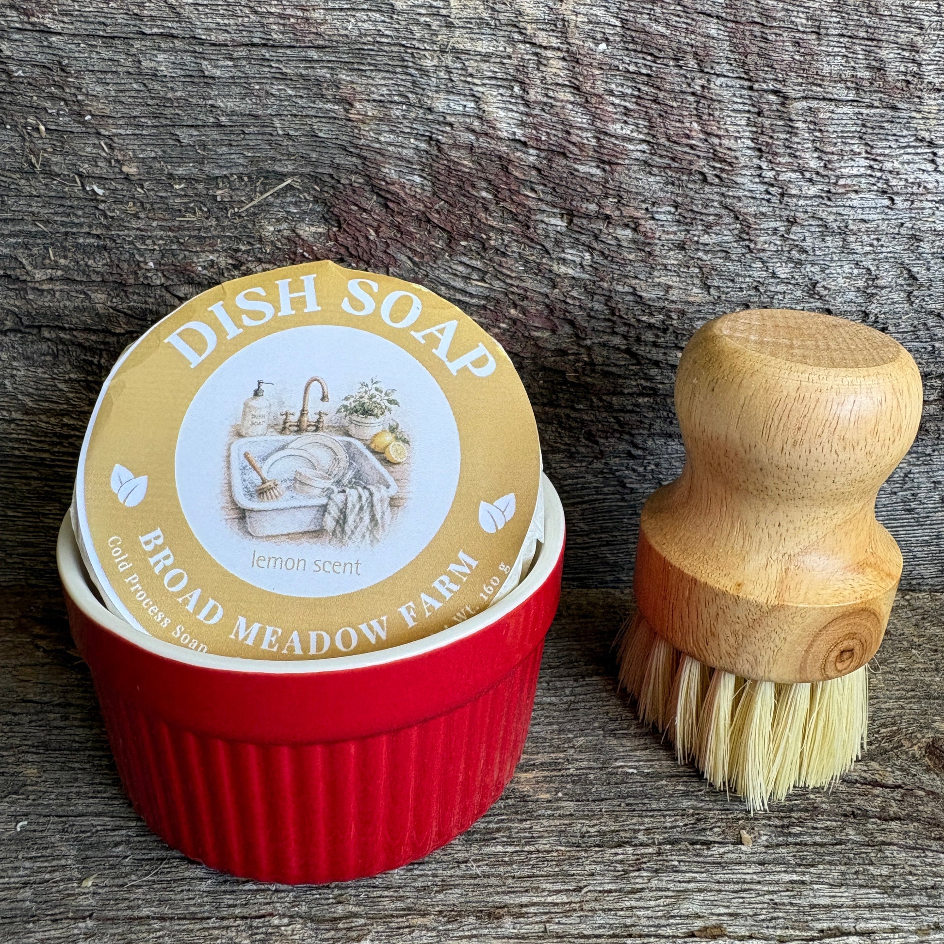 A red ribbed dish soap container with a round lid label reading 'DISH SOAP BROAD MEADOW FARM lemon scent,' shown next to a small wooden-handled brush with cream bristles on a rustic weathered wood surface.