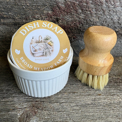 A small round dish soap tin labeled 'DISH SOAP BROAD MEADOW FARM lemon scent' sits in a white ribbed ramekin next to a wooden-handled cleaning brush on a rustic weathered wood surface.