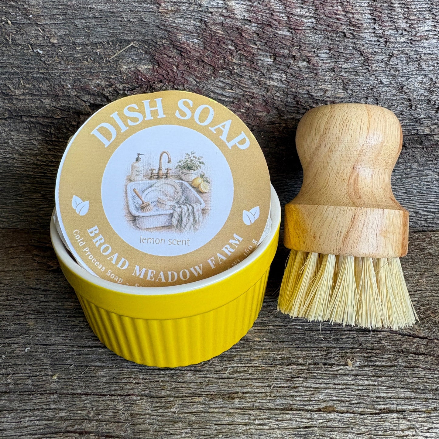 Yellow dish soap tin with a wooden-handled brush on a rustic wood background. The round lid reads 'DISH SOAP' and 'BROAD MEADOW FARM' with lemon imagery, and the brush has a light wood handle and pale bristles.