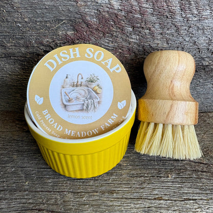 Yellow dish soap tin with a wooden-handled brush on a rustic wood background. The round lid reads 'DISH SOAP' and 'BROAD MEADOW FARM' with lemon imagery, and the brush has a light wood handle and pale bristles.