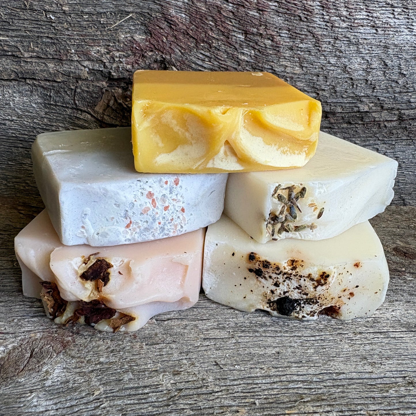 Five artisanal handmade soap bars stacked on a rustic weathered wood background. The soaps feature natural variations in color and embedded botanicals, including a prominent yellow marbled bar on top, a white bar with pink speckles, a pale pink bar with dark brown botanicals, and two cream-colored bars with dark charred-looking inclusions.