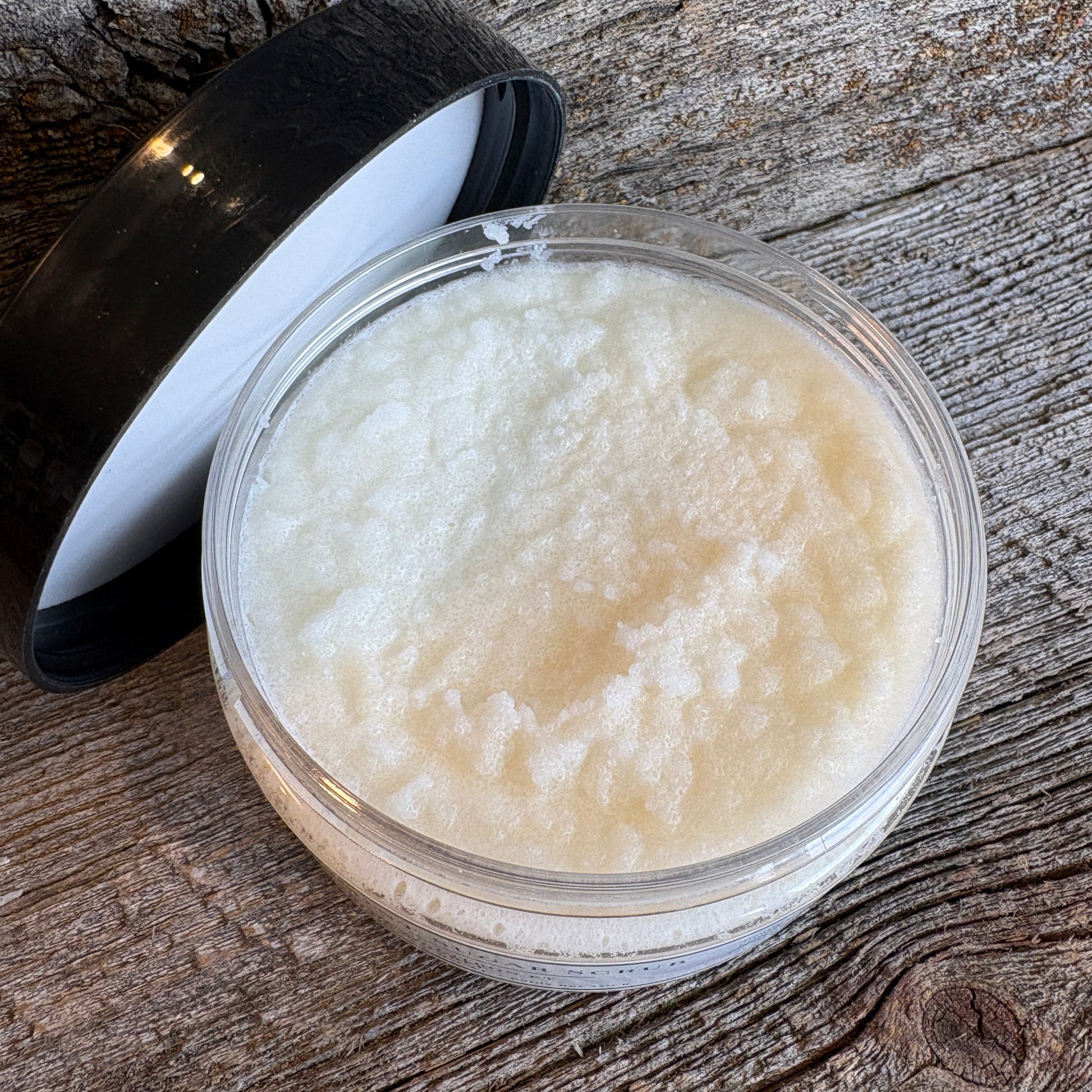 A clear round plastic jar filled with a thick, creamy off-white balm or scrub, shown with its black lid resting beside it on a rustic weathered wood surface.