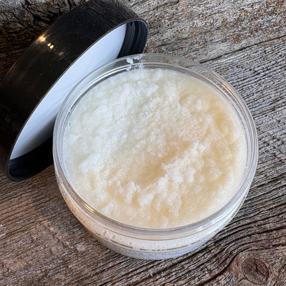 A clear round plastic jar filled with a thick, creamy off-white balm or scrub, shown with its black lid resting beside it on a rustic weathered wood surface.