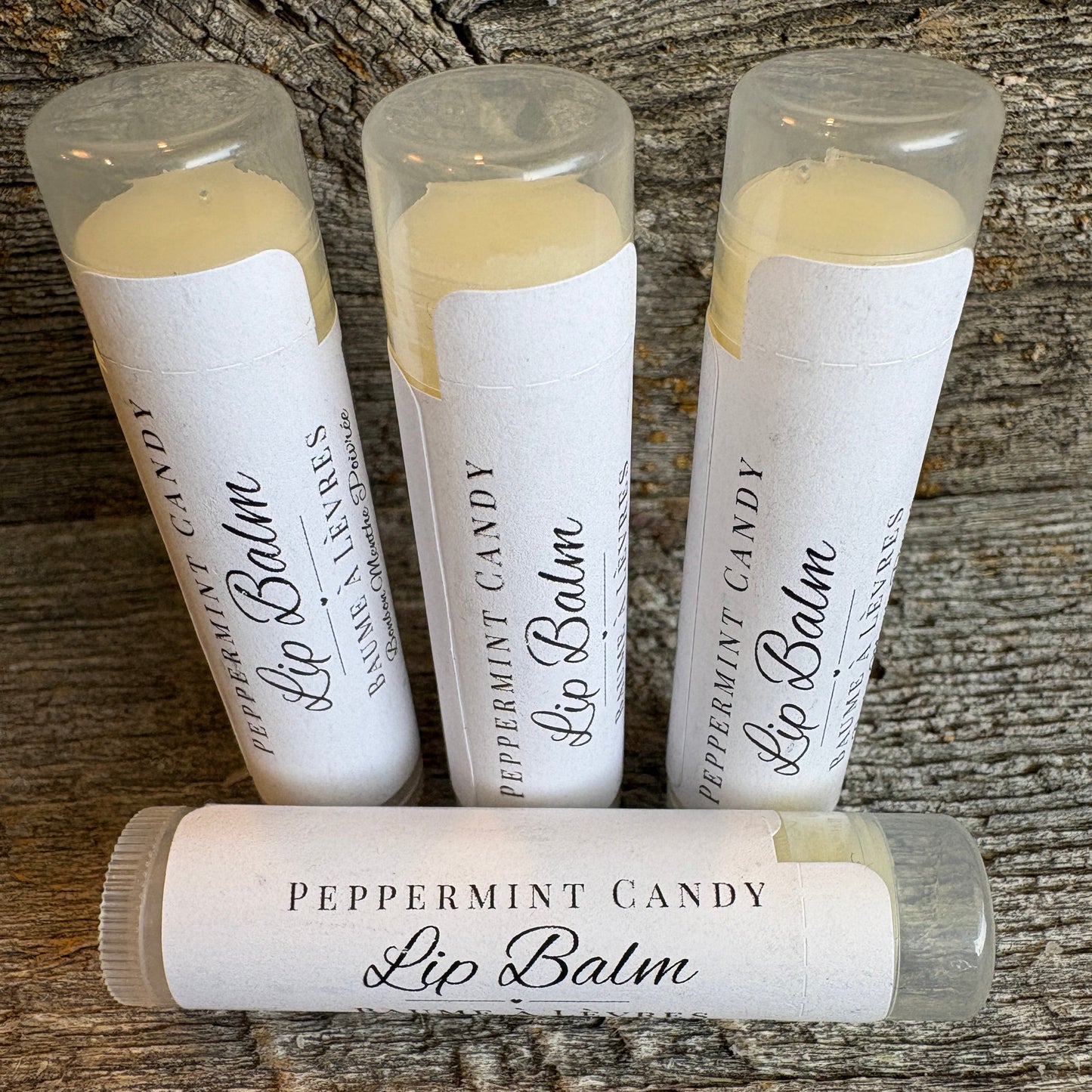 Four lip balm tubes labeled 'Peppermint Candy Lip Balm' arranged on a rustic wooden surface, with three standing upright and one lying flat in front.