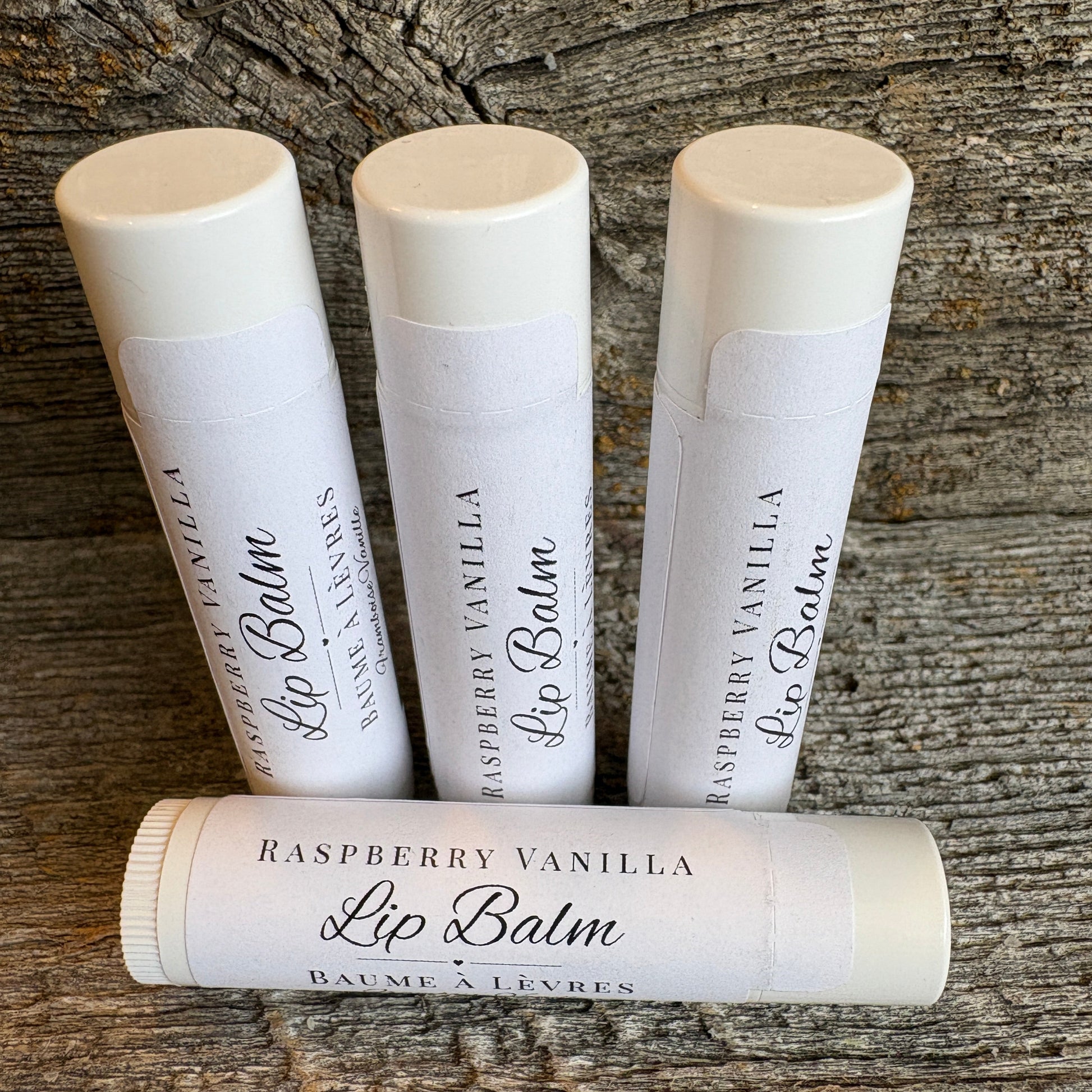 Four tubes of Raspberry Vanilla Lip Balm arranged on a rustic weathered wood surface, with three standing upright and one lying horizontally in the foreground.