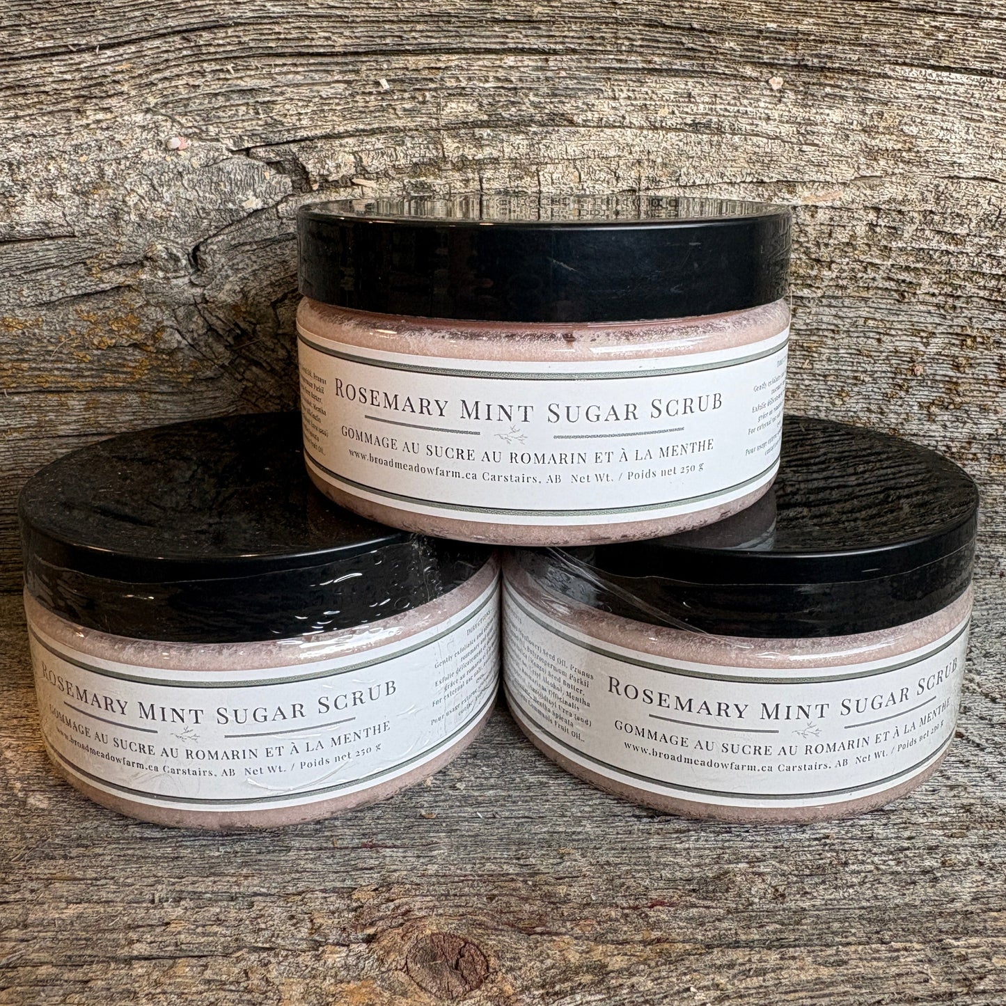 Three jars of Rosemary Mint Sugar Scrub stacked in a pyramid on a rustic wooden surface. The jars have black lids and white labels with black text.