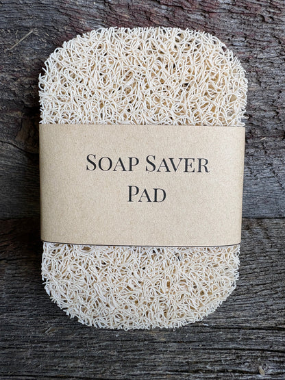 A synthetic fibre soap saver pad wrapped in a kraft paper band labeled 'SOAP SAVER PAD', placed on a dark rustic wooden surface.