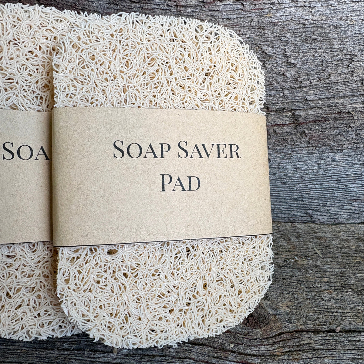 Two synthetic soap saver pads wrapped in kraft paper bands labeled 'SOAP SAVER PAD', displayed on a rustic weathered wood background.