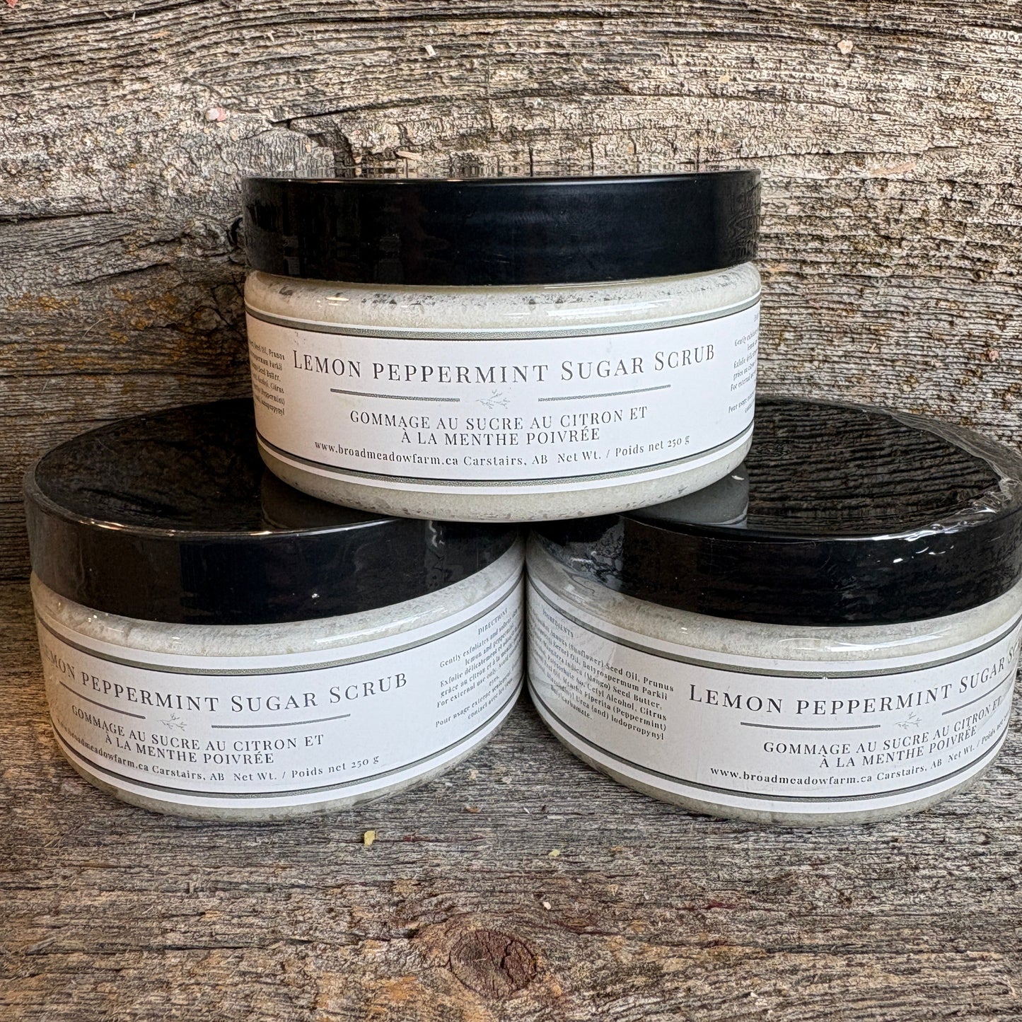 Three jars of Lemon Peppermint Sugar Scrub arranged in a pyramid on a rustic wooden surface. The white jars have black lids and white labels with black text.