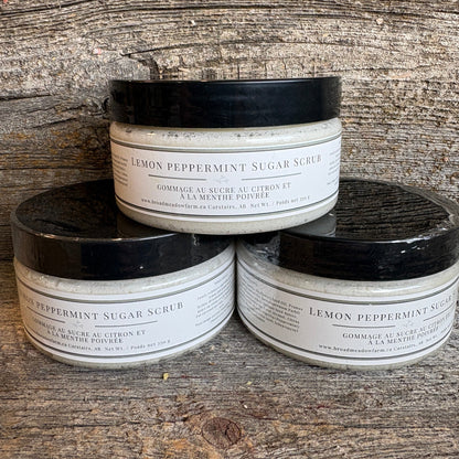 Three jars of Lemon Peppermint Sugar Scrub arranged in a pyramid on a rustic wooden surface. The white jars have black lids and white labels with black text.