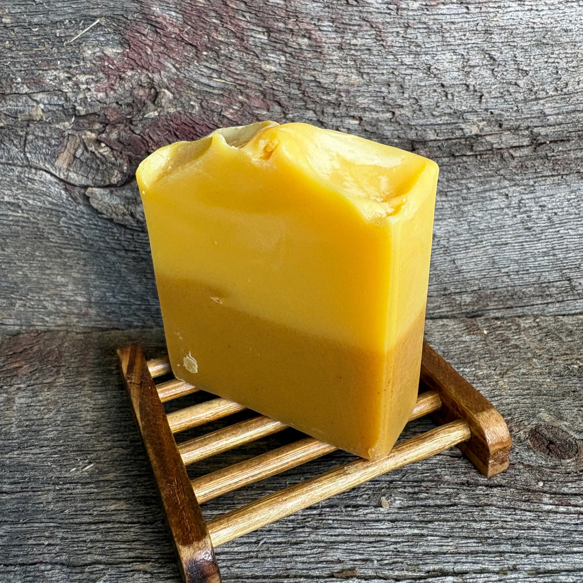 A handmade rectangular bar of yellow soap resting on a small wooden slatted stand against a rustic weathered wood background.