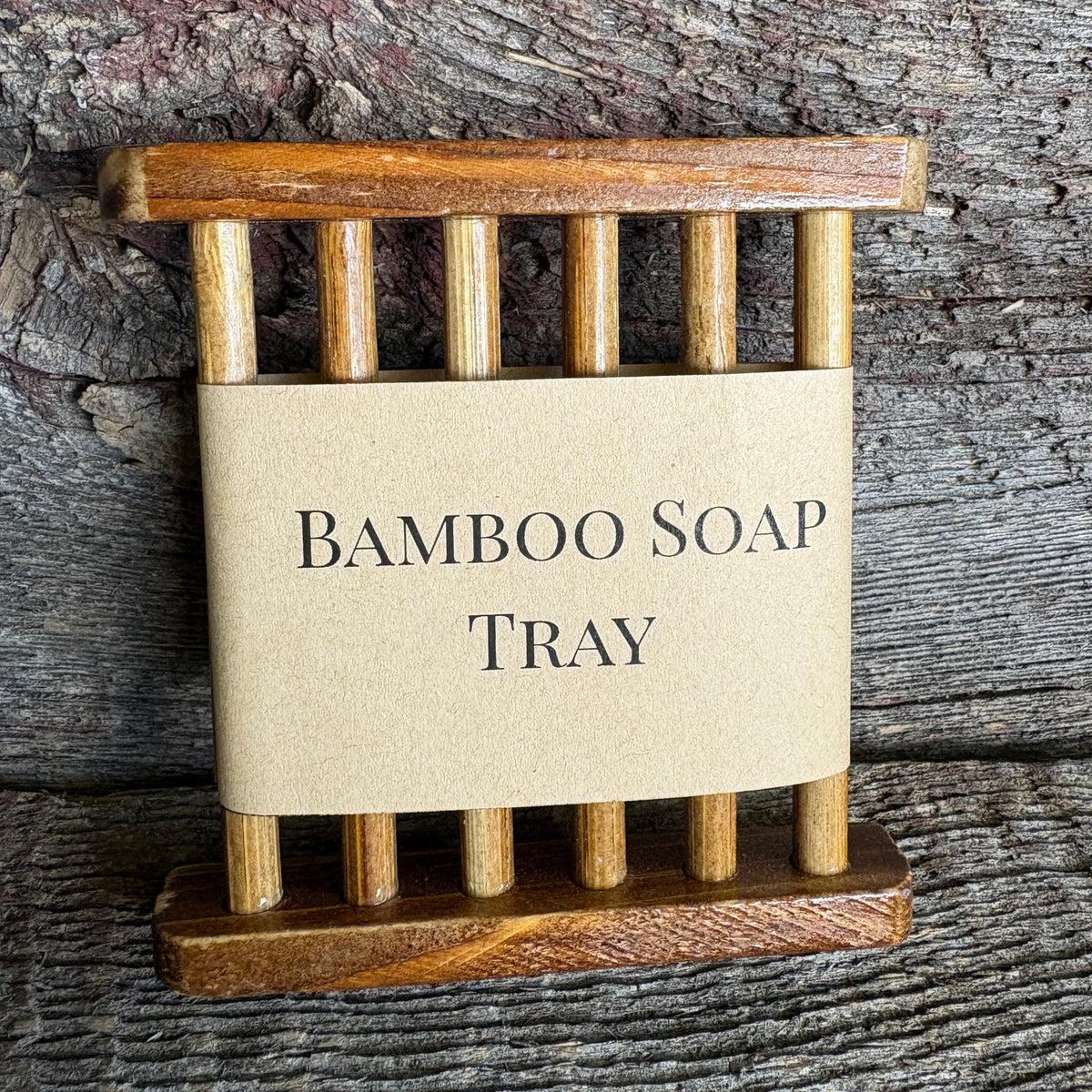 A small bamboo soap tray with a natural kraft paper label reading 'BAMBOO SOAP TRAY' in black serif text, photographed against a rustic weathered wood background.