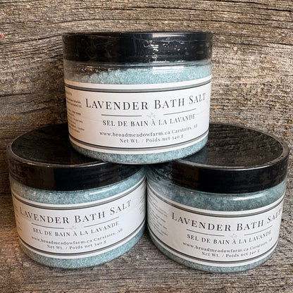 Three jars of Broadmeadow Farm Lavender Bath Salt stacked on a rustic wooden surface. The jars contain a light blue-green bath salt and are fitted with black lids and white labels.