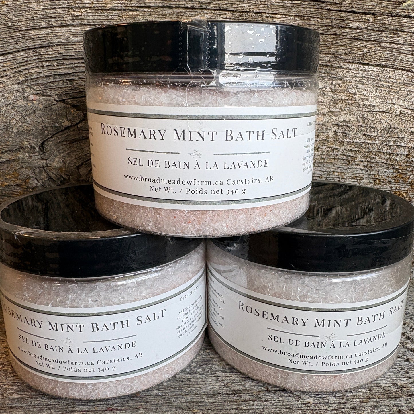 Three jars of Broadmeadow Farm Rosemary Mint Bath Salt stacked in a pyramid on a rustic wooden surface.