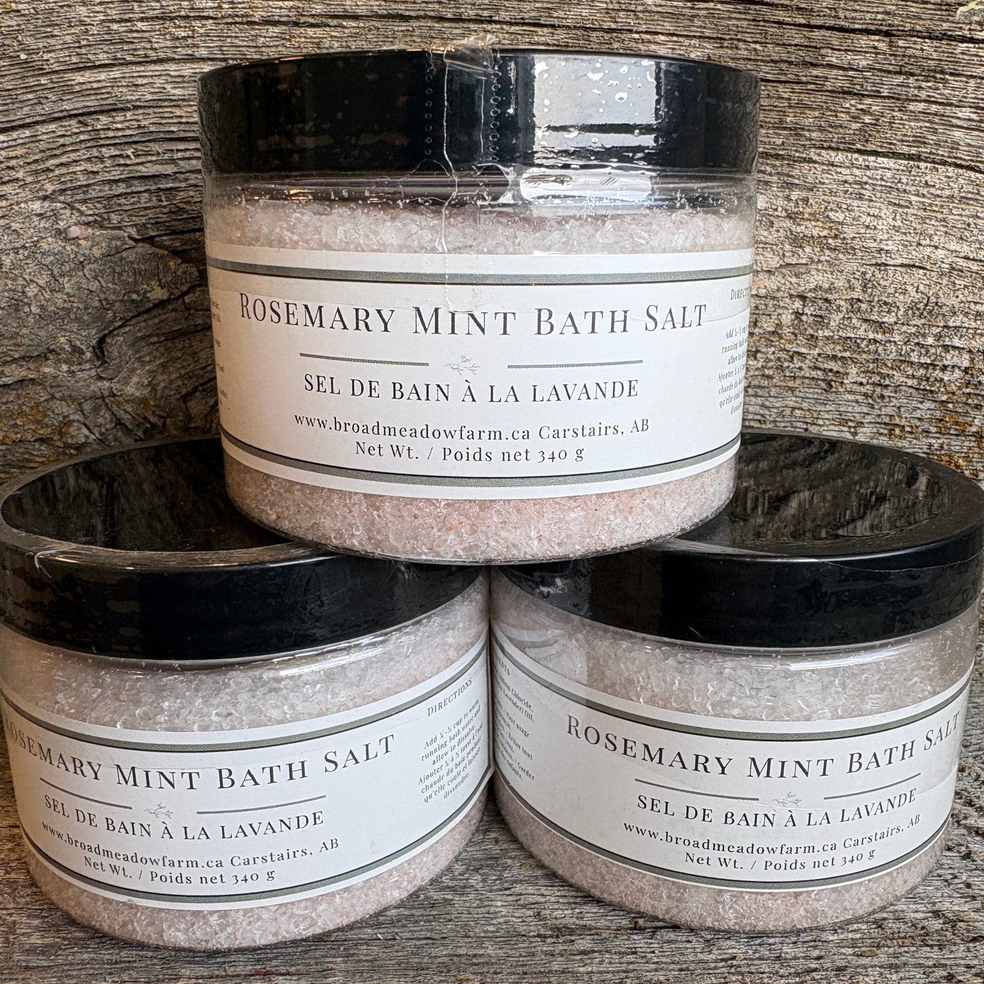 Three jars of Broadmeadow Farm Rosemary Mint Bath Salt stacked in a pyramid on a rustic wooden surface.