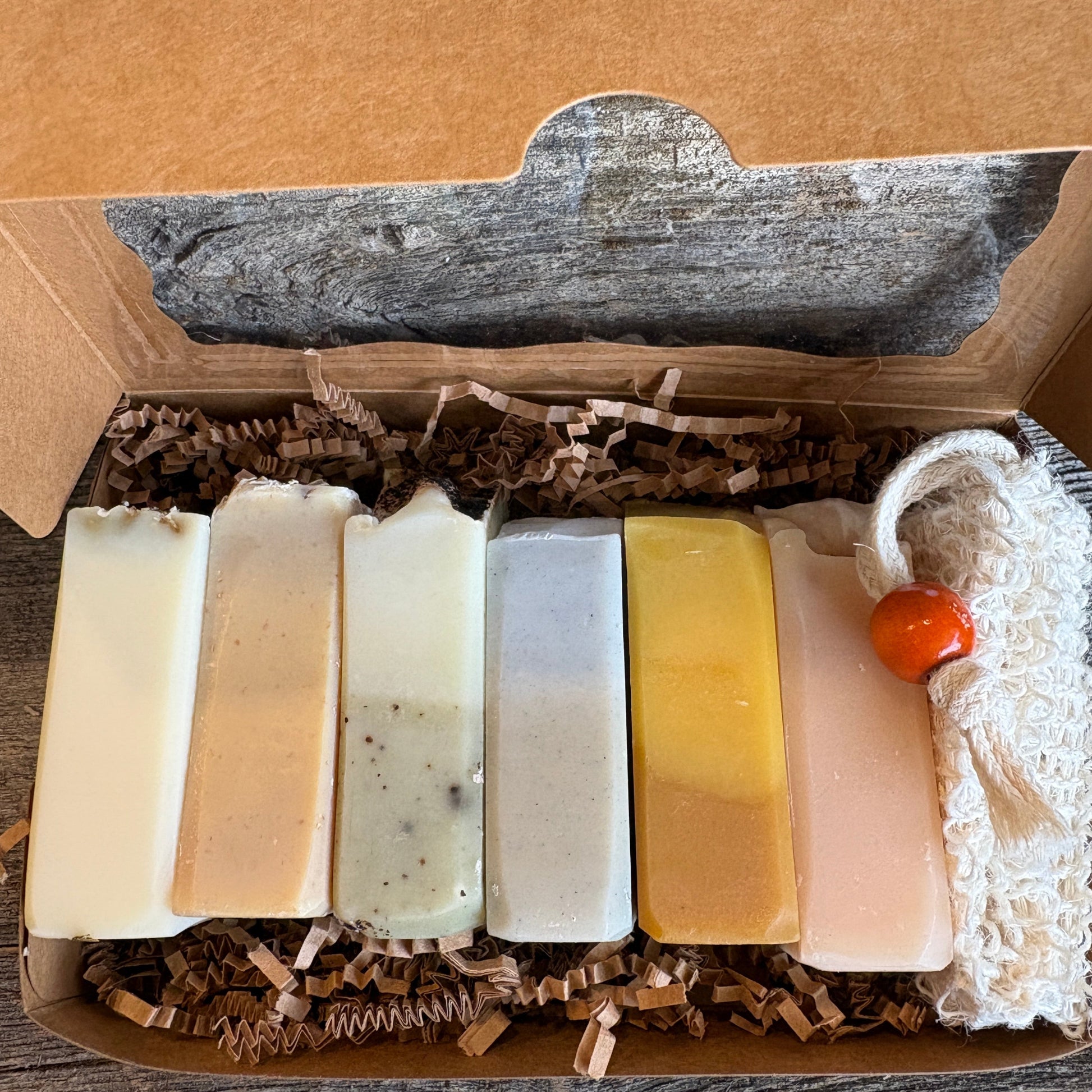 An open kraft paper box filled with shredded paper packaging, holding six rectangular bars of soap in different natural tones, with a white textured cloth and small orange bead resting on the right side, set on a rustic wooden surface.