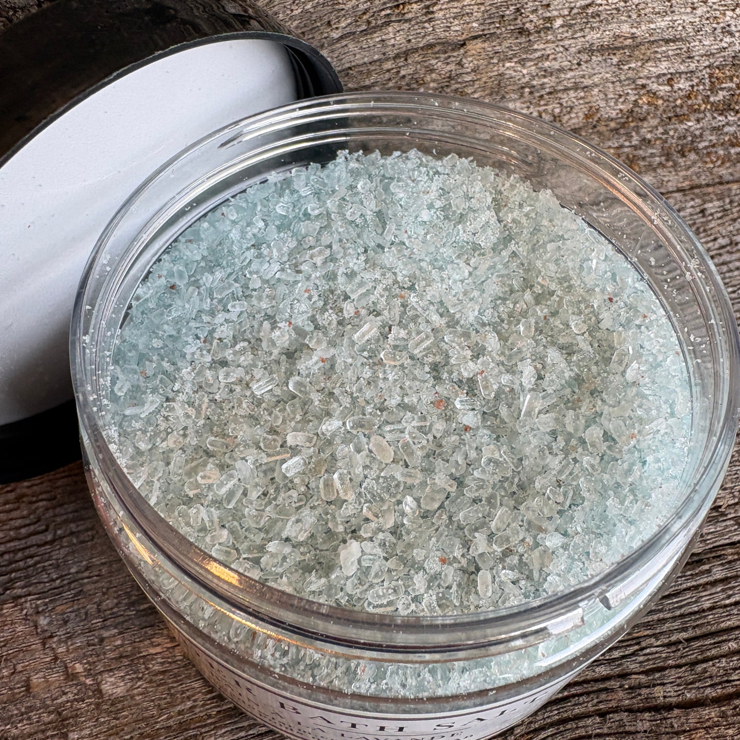 A clear round jar filled with pale icy blue-green crystal bath salts, shown on a rustic weathered wood surface with a white and black lid resting beside it.