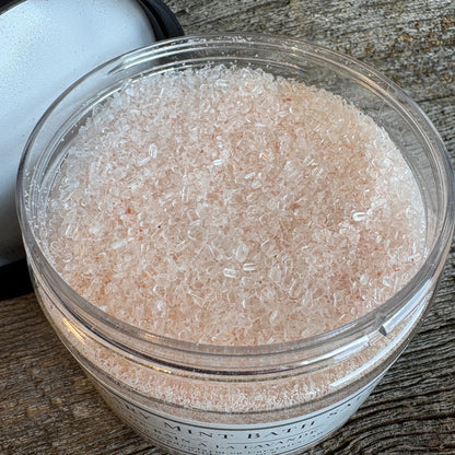 A clear plastic jar filled with pale pink bath salts, shown on a rustic weathered wood surface with the black-and-white lid partially visible behind it.