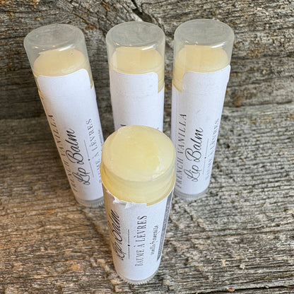 Four tubes of lip balm with white labels and clear caps, showing pale yellow balm inside, arranged on a rustic weathered wood surface. The front tube is open, showing the balm, while the others are closed.