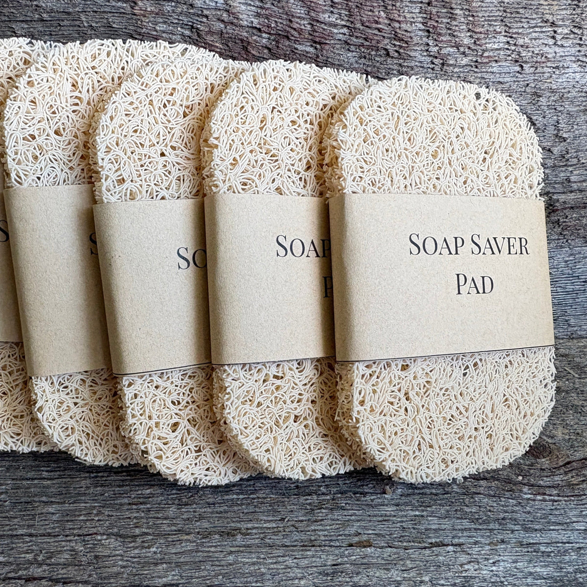 Five synthetic loofah soap saver pads wrapped in kraft paper bands, arranged in a fan on a rustic weathered wood background.