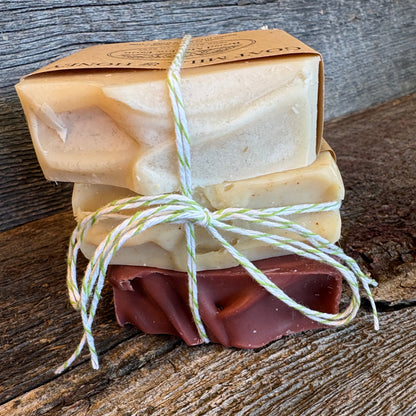 3 handmade soap bars tied with twine, one in natural cream and one in dark brown, displayed on a rustic wooden surface next to a kraft paper box.