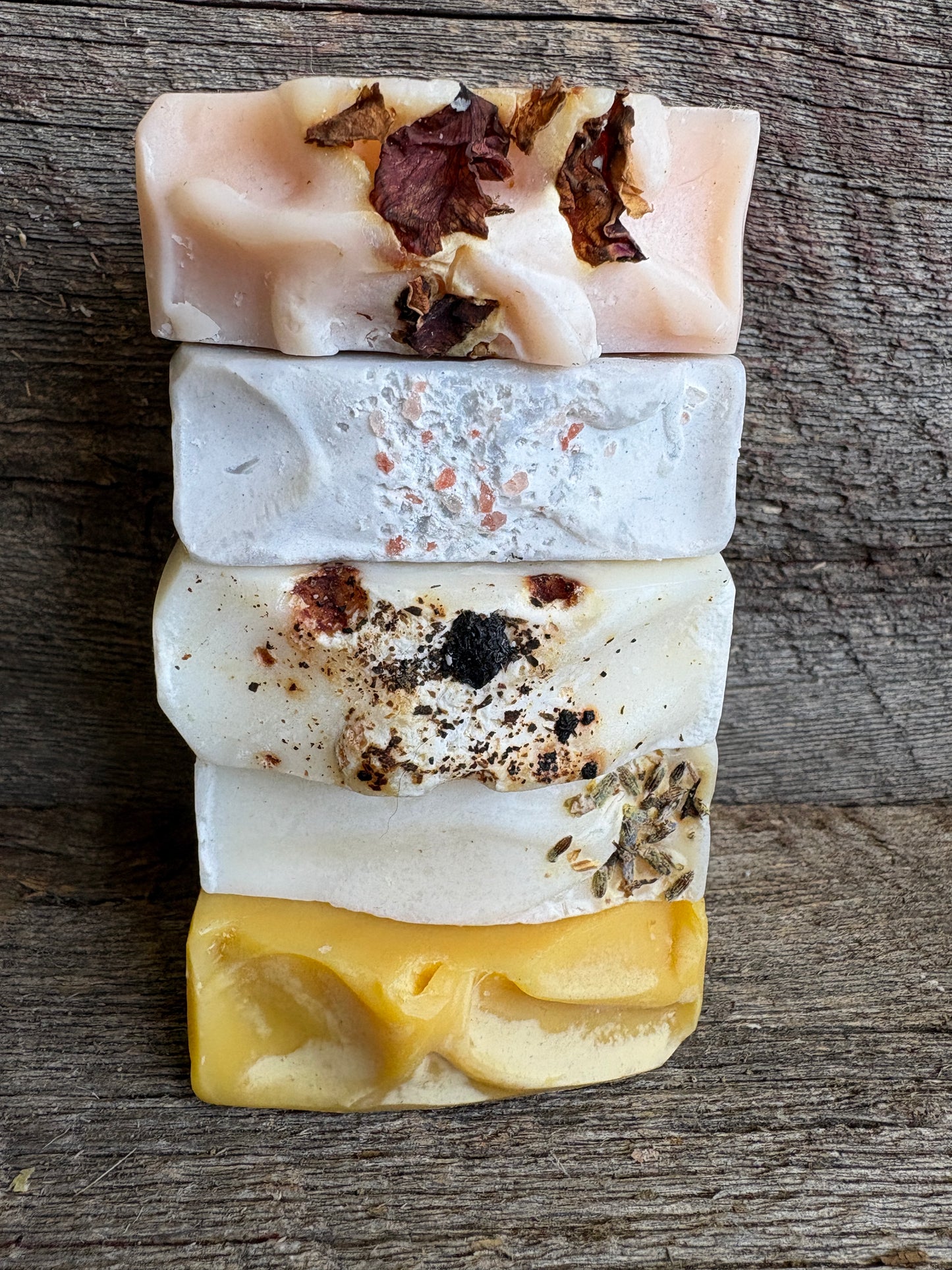 Five stacked handmade soap bars in different colors and textures, including pink with dried flowers, white with pink flecks, white with dark specks, white with dried herbs, and yellow, arranged vertically on a rustic wooden surface.